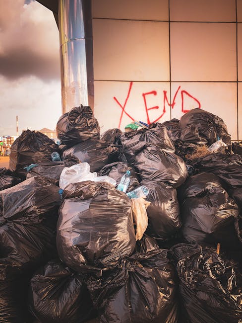 A large pile of black plastic rubbish bags, some torn or partially open, stacked against a modern building with smooth, light-colored tiled exterior panels. The bags contain various waste materials, w