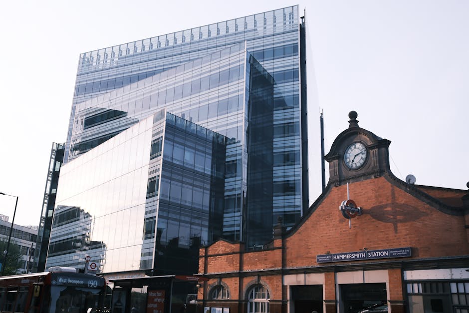 The image depicts a modern, multi-story office building with a glass facade reflecting the surrounding structures and overcast sky, situated behind an older brick building that features Victorian arch